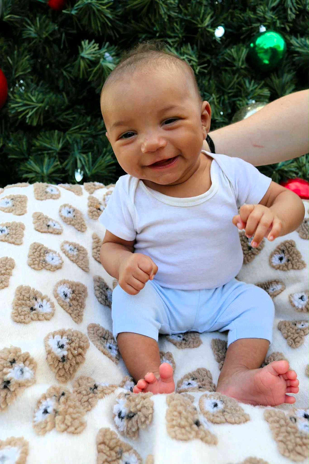 Kyàire joined the competition — help win amazing prizes! baby, smiling, blanket, teddy_bear_pattern, christmas_tree, ornaments, greenery, infant, cute, happy, white_shirt, blue_pants, toes, hand, face, outdoor, festive, holiday, person, sitting