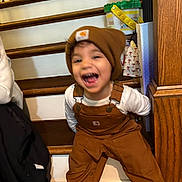 Kaige is registered to the contest to win money with this photo: beanie, brown_overalls, cabinet, casual, child, clothing, cute, footwear, happy, indoor, laughing, person, playful, smiling, stairs, toddler, toy_bucket, white_shirt, wood_floor, wooden_stairs