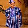 child, traditional_clothing, hat, beads, blue, maroon, striped, standing, portrait, indoor, decor, plant, woven_mat, serious_expression, black_shoes, cultural, fashion, person, young, accessory