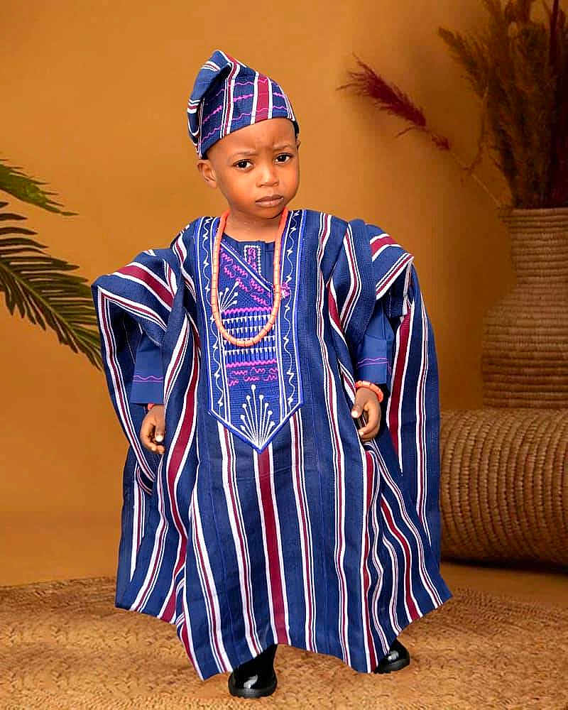 Abraham joined the competition — help win amazing prizes! child, traditional_clothing, hat, beads, blue, maroon, striped, standing, portrait, indoor, decor, plant, woven_mat, serious_expression, black_shoes, cultural, fashion, person, young, accessory