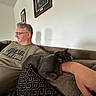 man, cat, couch, arm, glasses, gray_cat, relaxed, indoor, wall_art, cushion, pattern, beard, short_hair, casual_clothing, resting, home, seated, portrait, living_room, comfort
