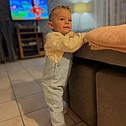 Maëlio participe au concours pour gagner de l'argent avec cette photo : toddler, child, indoor, living_room, overalls, sneakers, couch, blanket, television, cartoon, tile_floor, lamp, curtains, furniture, person, cute, standing, casual_clothing, home, curious