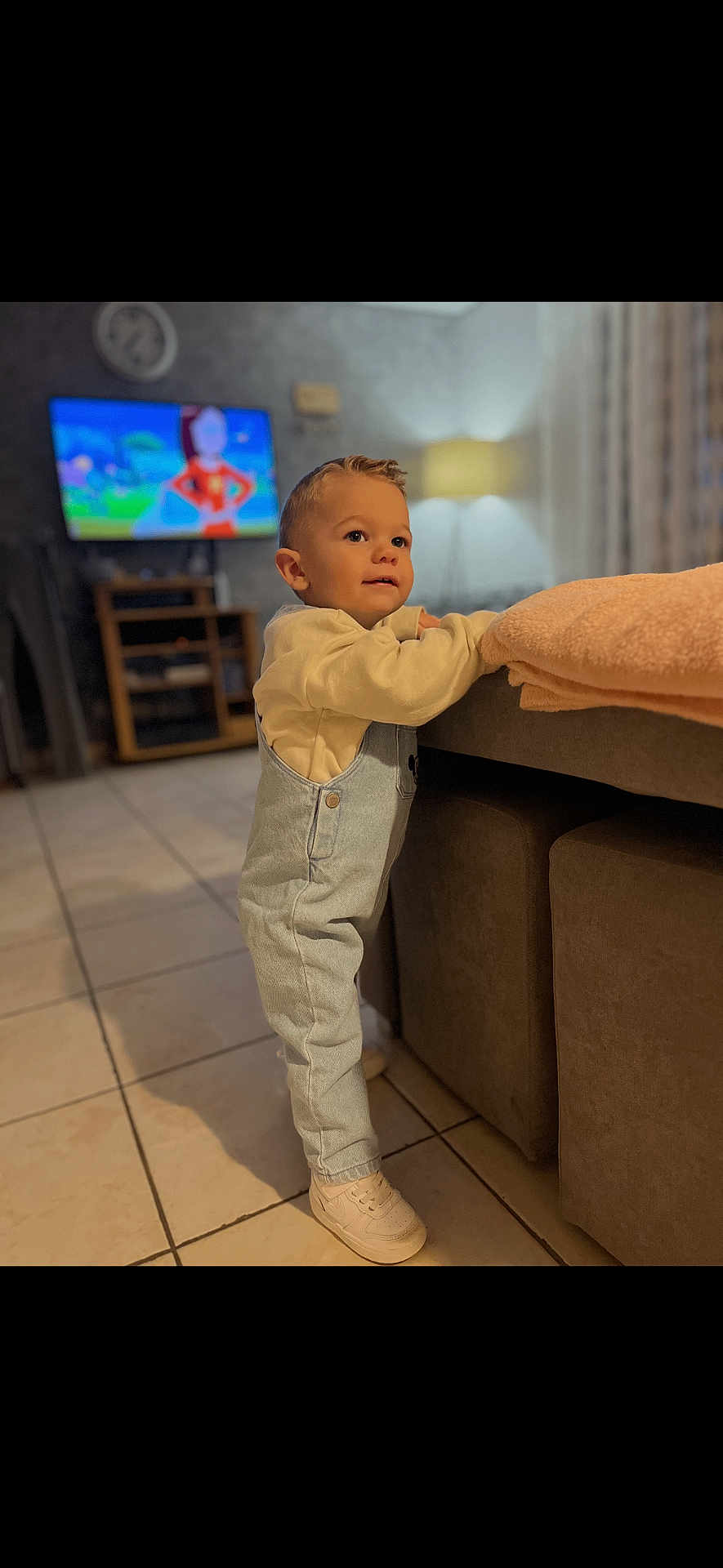 Maëlio participe au concours pour gagner de l'argent avec cette photo : toddler, child, indoor, living_room, overalls, sneakers, couch, blanket, television, cartoon, tile_floor, lamp, curtains, furniture, person, cute, standing, casual_clothing, home, curious