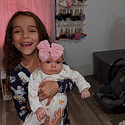 Haisley is registered to the contest to win money with this photo: child, baby, pajamas, pink_bow, smiling, holding, indoor, wooden_floor, curtains, hanger, clothing, car_seat, floor, person, face, happy, infant, room, decor, footwear