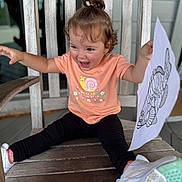 Athena is registered to the contest to win money with this photo: child, clothing, cute, drawing, fish, happy, indoor, joyful, leggings, paper, playful, pony_tail, portrait, shoes, sitting, smiling, toddler, tshirt, wood, wooden_chair