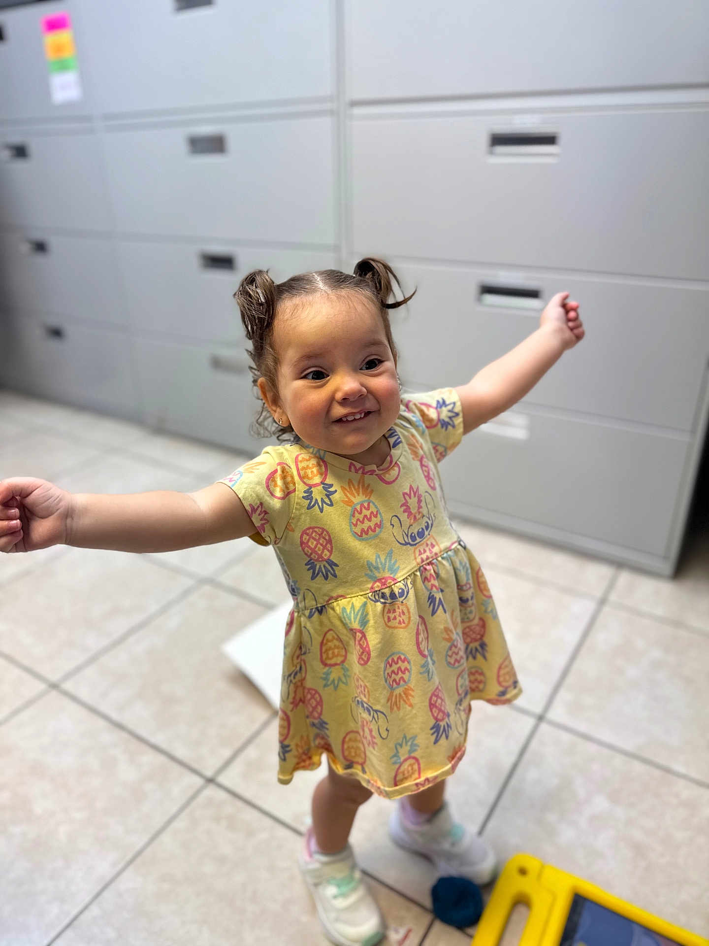 Athena joined the competition — help win amazing prizes! toddler, child, smiling, yellow_dress, pineapple_pattern, pigtails, indoor, tile_floor, filing_cabinet, toy, sneakers, happy, arms_outstretched, person, young_child, cute, playful, small_toy, floor, expression