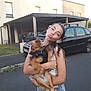 girl, child, dog, pet, holding, street, car, house, carport, summer, casual_clothing, denim_shorts, white_tank_top, playful, portrait, outdoor, animal, young, fun, daytime