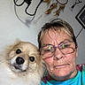 dog, person, glasses, tie_dye_shirt, selfie, indoor, dreamcatcher, wall_decor, close_up, pet, fluffy_dog, face, portrait, casual, adult, home, cute, looking_at_camera, brown_hair, eyewear