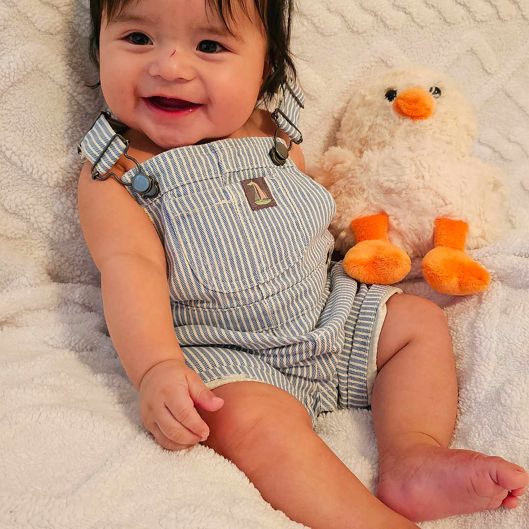 Hannibal is registered to the contest to win money with this photo: baby, blanket, child, cozy, cute, duck, face, foot, furniture, hand, happy, indoor, infant, playful, plush_toy, portrait, sitting, smiling, soft_toy, striped_overalls