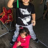 children, child, toddler, sibling, standing, sitting, indoors, portrait, smiling, toy, green_toy, red_outfit, shark_shirt, curly_hair, wood_floor, folding_chair, playtime, family, casual_clothing, home