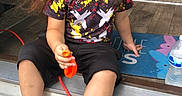 Andy is registered to the contest to win money with this photo: child, toddler, portrait, sitting, outdoor, doorstep, curly_hair, long_hair, facial_expression, looking_at_camera, casual_clothing, colorful_shirt, black_shorts, sneakers, orange_toy, water_bottle, welcome_mat, house_entrance, living_room, daylight