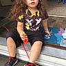 child, toddler, portrait, sitting, outdoor, doorstep, curly_hair, long_hair, facial_expression, looking_at_camera, casual_clothing, colorful_shirt, black_shorts, sneakers, orange_toy, water_bottle, welcome_mat, house_entrance, living_room, daylight