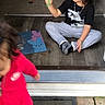 child, children, toddler, pirate_hat, sitting, cross_legged, toy, green_toy, red_clothing, black_shirt, gray_pants, shoes, doorway, porch, wooden_floor, toys, chalkboard, indoor, outdoor, candid