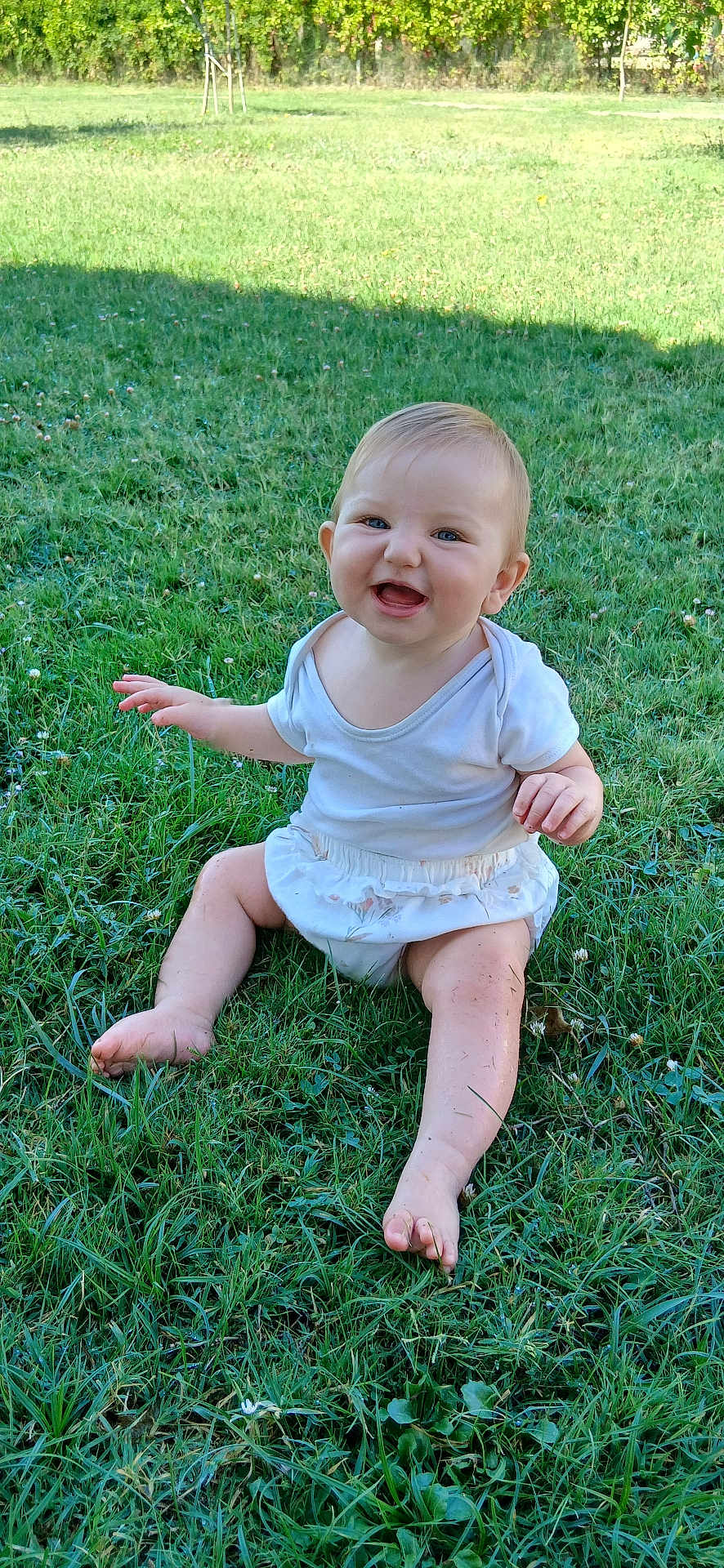 Lina-Maria participe au concours pour gagner de l'argent avec cette photo : baby, child, grass, outdoor, smiling, happy, sitting, nature, greenery, sunlight, skirt, white_top, barefoot, cute, infant, playful, person, head, face, summer