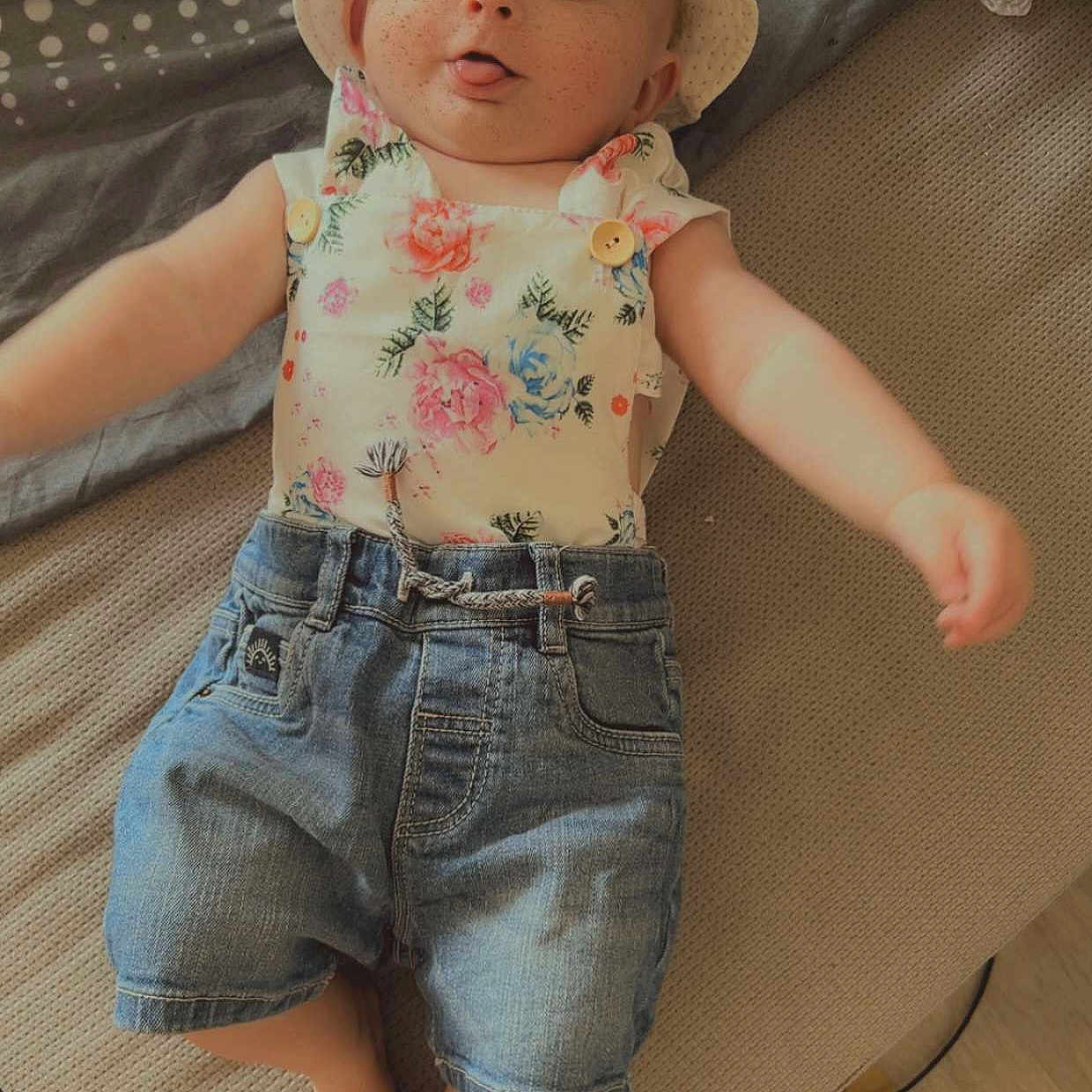 Jade participe au concours pour gagner de l'argent avec cette photo : baby, barefoot, bed, child, clothing, cute, denim_shorts, fabric, floral_top, hat, headwear, indoor, infant, lying_down, person, playful, polka_dot, skin, smartphone, tongue_out