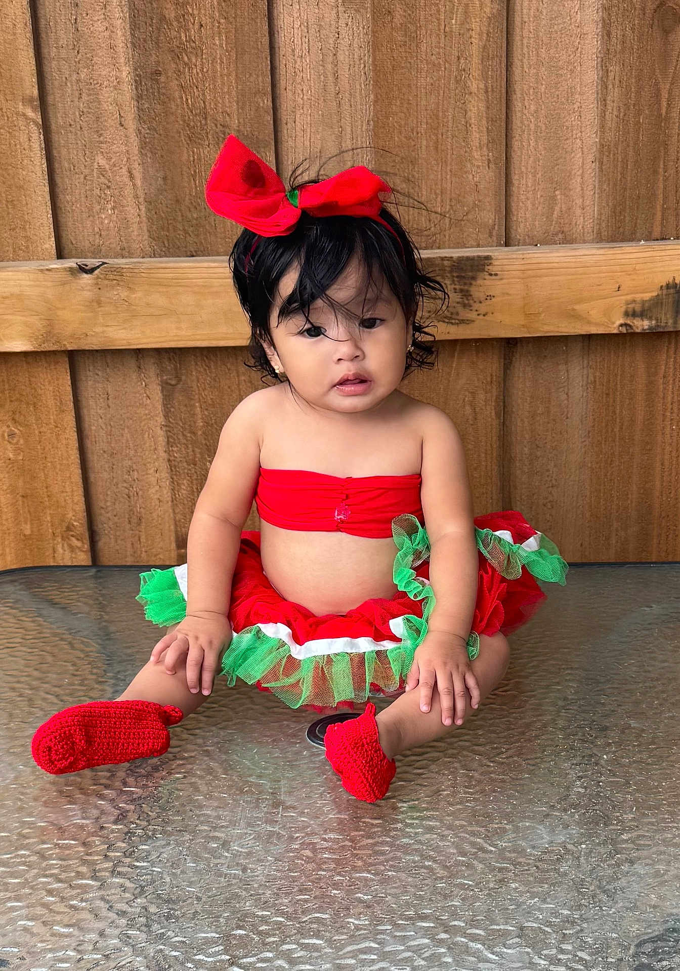 Evelyn is registered to the contest to win money with this photo: toddler, child, red_bow, tutu_skirt, red_booties, glass_table, wooden_fence, curly_hair, sitting, outdoor, festive_clothing, cute, portrait, baby, headband, colorful_clothing, young_child, curious_expression, casual_pose, daylight