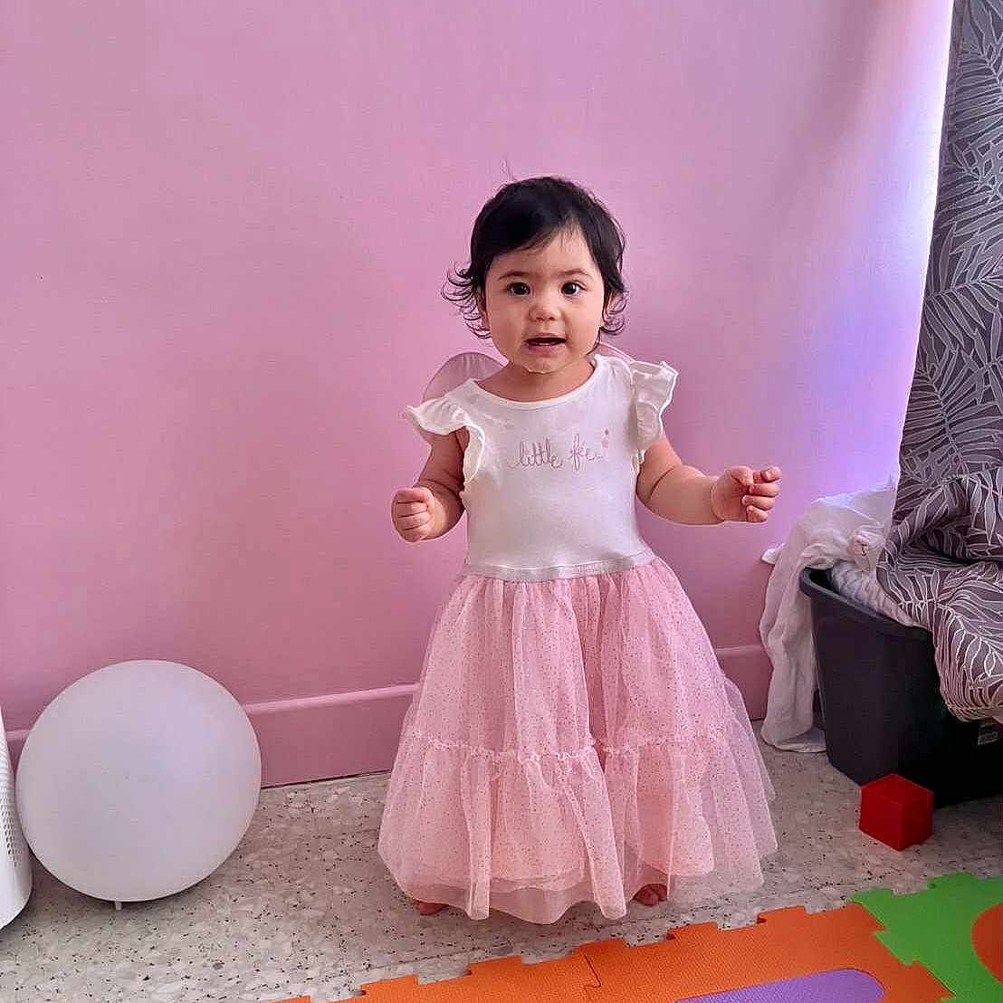 Estella a rejoint le concours — aidez-le/la à gagner de superbes lots ! baby, child, colorful_mats, curtain, cute, floor, girl, happy, home, indoor, person, pink_dress, play, playroom, smiling, standing, toddler, toy_block, wall, young_child