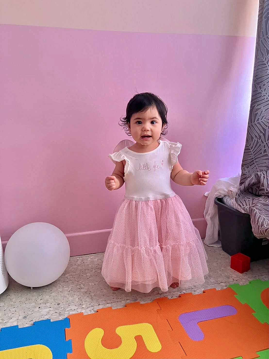 Estella a rejoint le concours — aidez-le/la à gagner de superbes lots ! toddler, child, pink_dress, standing, colorful_mats, indoor, playroom, curtain, toy_block, floor, young_child, smiling, person, baby, cute, wall, home, play, happy, girl
