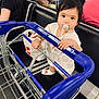 Estella participe au concours pour gagner de l'argent avec cette photo : toddler, child, pacifier, shopping_cart, airport, waiting_area, people, seated, luggage, blue_handle, white_clothing, black_hair, socks, metal_cart, bag, floor, bench, adult, phone, leopard_print