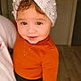 toddler, child, portrait, smile, headband, knitwear, pearls, brown_eyes, orange_clothing, necklace, indoor, person, cute, young_child, fashion, warm_colors, soft_lighting, happy, closeup, holding_hand