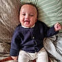baby, smiling, sitting, navy_sweater, white_pants, pillow, blanket, cozy, indoors, cute, child, happy, face, person, infant, soft, comfort, home, portrait, warm
