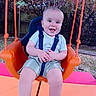 Lorenzo is registered to the contest to win money with this photo: baby, child, daylight, fence, fun, grass, happy, harness, orange, outdoor, person, play, playground, seat, shorts, smiling, striped_shirt, summer, swing, trees
