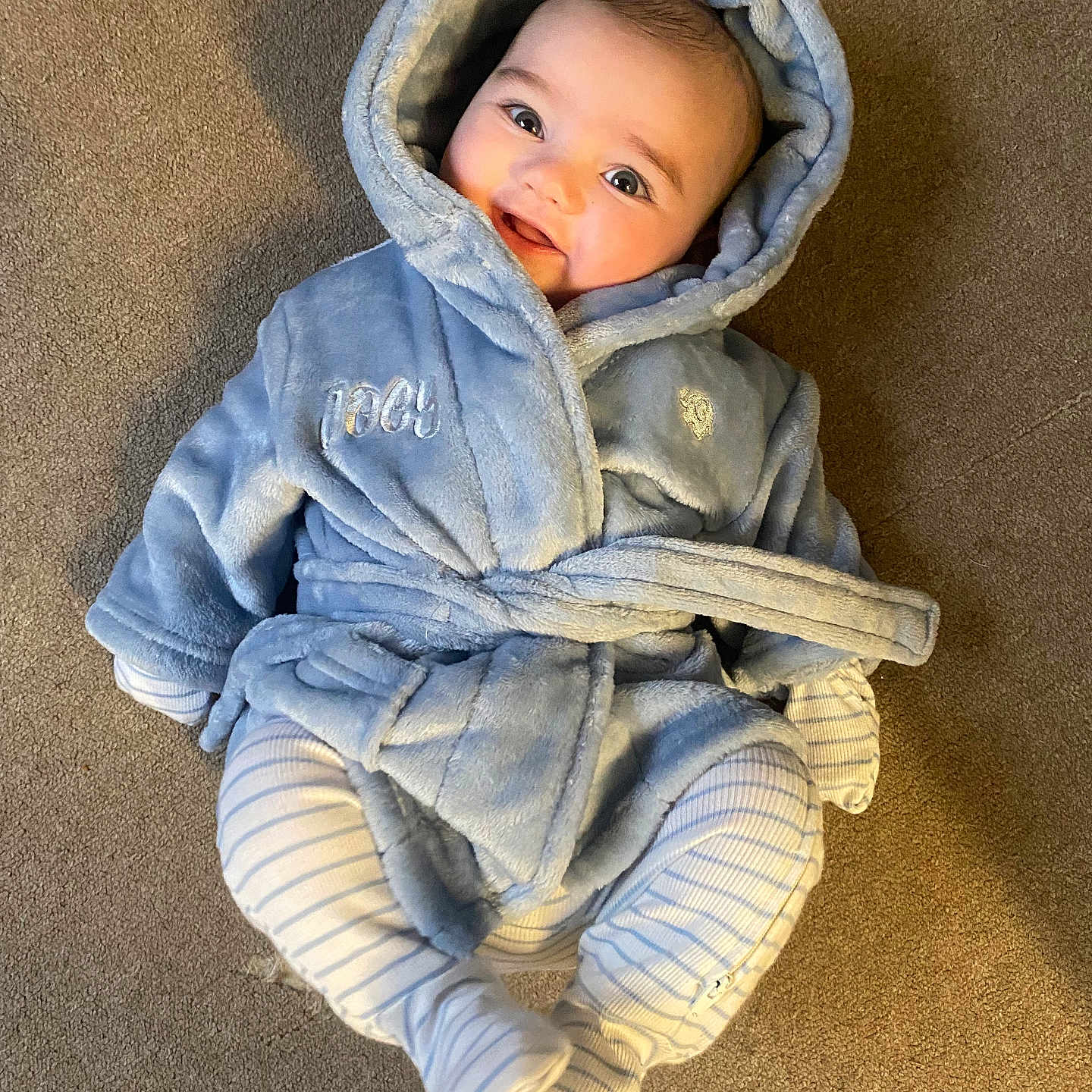 Joey is registered to the contest to win money with this photo: apparel, baby, blue_robe, carpet, child, cozy, cute, face, happy, hooded_clothing, indoors, infant, lying_down, person, portrait, smiling, soft_texture, striped_leggings, warm_colors, young_child