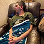 man, kitten, cat, blanket, chair, leather_chair, glasses, casual_clothing, indoor, relaxing, seated, pet, animal, comfort, resting, brown_furniture, short_hair, beard, hands, cozy