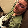 man, glasses, sleeping, kitten, cat, blanket, couch, indoor, relaxing, beard, green_shirt, chain_necklace, pet, snuggling, comfort, resting, fur, paw, closeup, human