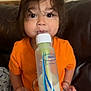 baby_bottle, blanket, casual, child, couch, cozy, curious, expression, eyes, face, feeding, hair, indoors, milk, milk_bottle, orange_shirt, person, sitting, toddler, young_child