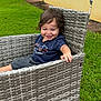 child, toddler, smiling, happy, outdoor, grass, chair, woven_chair, blue_shirt, shorts, person, playful, curly_hair, summer, relaxing, daylight, greenery, yellow_wall, casual_clothing, candid