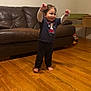 toddler, child, barefoot, standing, arms_up, smiling, indoor, wooden_floor, leather_couch, toy, desk, clothing, happy, person, home, furniture, young_child, dark_clothes, living_room, expression