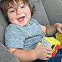 blue_shorts, button, car_interior, car_seat, child, cute, face, gray_shirt, hair, hand, happy, playful, seat, seat_belt, short_sleeve_shirt, smiling, toddler, toy, yellow_toy, young_child