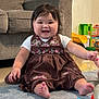 baby, child, smiling, sitting, dress, brown_dress, embroidery, indoor, carpet, living_room, toy, foot, hand, happy, cute, person, face, hair, couch, floor
