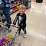 toddler, child, shopping_cart, grocery_store, skeleton_outfit, black_clothing, white_shoes, floor_tiles, snacks, candy, adult, jeans, plaid_shirt, smile, shopping, aisle, shelves, products, indoor, person