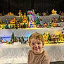 Milo is registered to the contest to win money with this photo: child, smile, blonde_hair, short_sleeve_shirt, jeans, miniature_village, christmas_tree, holiday_lights, festive, decorations, toys, indoor, seasonal, happy, portrait, seated, background, warm_lighting, winter_theme, playful