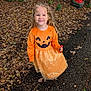 child, girl, smile, orange_dress, pumpkin_face, lollipop, curly_hair, autumn_leaves, outdoor, pathway, tree, nature, fall, happy, cute, seasonal, costume, playful, daylight, young