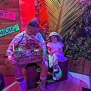 Ivy-violet is registered to the contest to win money with this photo: child, man, dinosaur_puppet, crate, wooden_box, white_dress, safari_hat, boots, neon_lights, tropical_decor, palm_leaves, wooden_wall, flooring, face_paint, indoor, people, smile, toy, fun, posing