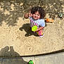 Logann is registered to the contest to win money with this photo: ball, child, colorful_clothing, curly_hair, daytime, footwear, hand_gesture, looking_up, outdoor, person, plaid_pants, play_area, playtime, sand, shadow, smiling, sneakers, sunlight, toddler, white_shirt