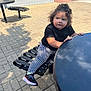 bench, black_shirt, building, car, casual_clothing, child, curious_expression, curly_hair, daytime, houndstooth_leggings, outdoor, person, shadow, sidewalk, sneakers, street, sunny, table, toddler, window