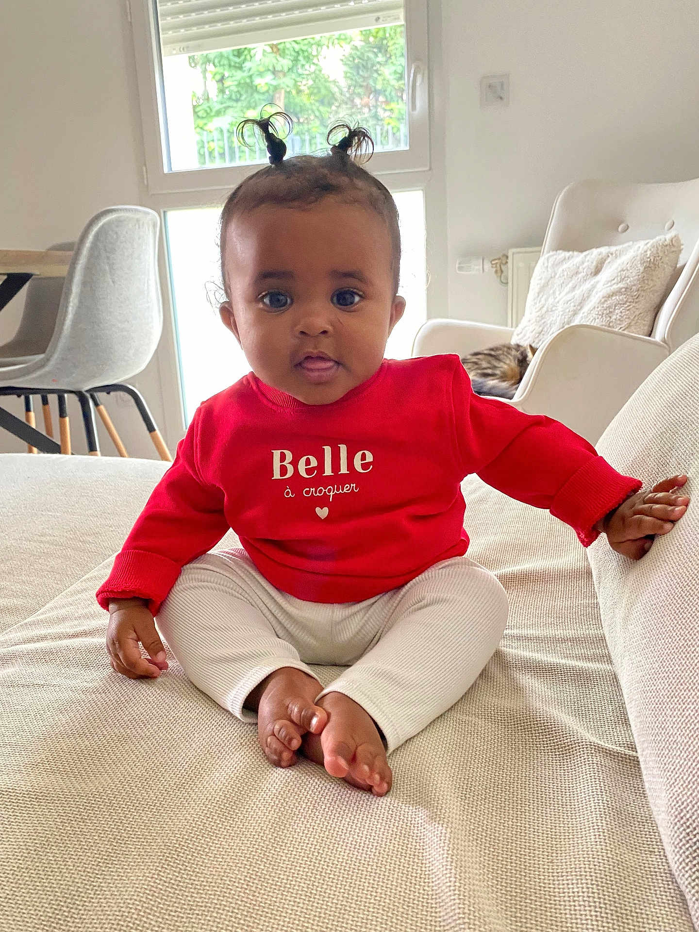 Haneya a rejoint le concours — aidez-le/la à gagner de superbes lots ! baby, child, indoor, couch, red_sweater, white_pants, pony_tail, cute, portrait, sitting, living_room, window, natural_light, furniture, pillow, floor, table, chair, person, home