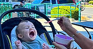 Thayer joined the competition — help win amazing prizes! baby, infant, child, yawning, sleepy, car_seat, spoon, feeding, adult, hands, outdoor, shopping_center, parking_lot, cars, bicycle, trees, sunlight, blue_sky, casual_photo, portrait