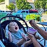 baby, infant, child, yawning, sleepy, car_seat, spoon, feeding, adult, hands, outdoor, shopping_center, parking_lot, cars, bicycle, trees, sunlight, blue_sky, casual_photo, portrait