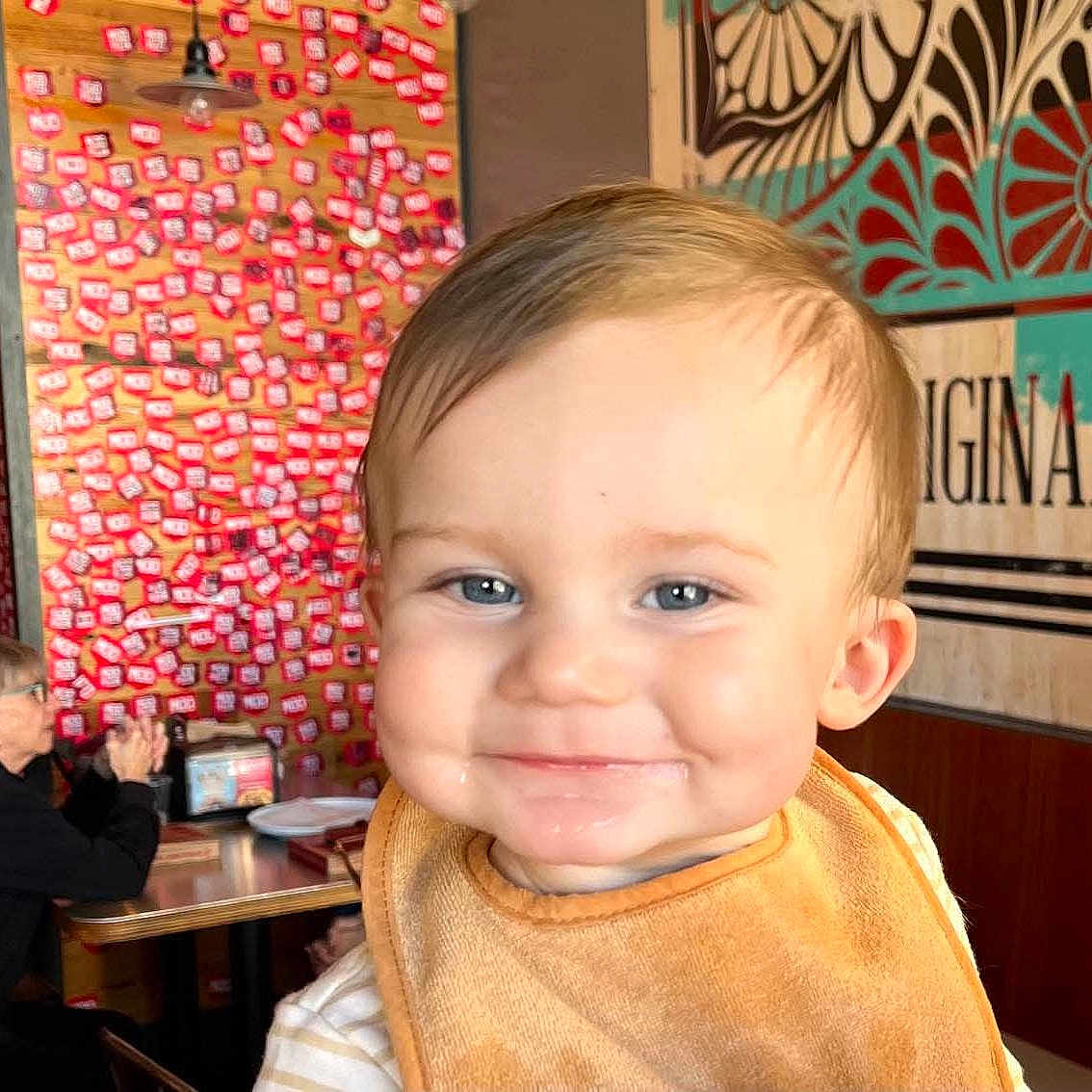Thayer joined the competition — help win amazing prizes! baby, child, toddler, smiling, portrait, face, close_up, indoor, restaurant, bib, orange, blond_hair, blue_eyes, seated, background_person, wall_art, colorful, table, casual, photography