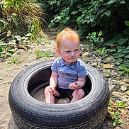 Oliver joined the competition — help win amazing prizes! baby, child, toddler, red_hair, face, sitting, tire, outdoor, garden, greenery, plants, rocks, pebbles, sunlight, portrait, infant, shirt, shorts, curious, playful