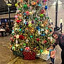 christmas_tree, ornaments, lights, toddler, child, elderly_woman, indoor, holiday, decorations, presents, people, scarf, sweater, floor, table, chairs, festive, celebration, smile, curious