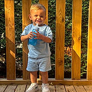 Liam participe au concours pour gagner de l'argent avec cette photo : toddler, child, smiling, outdoor, wooden_deck, greenery, sunlight, blue_shirt, shorts, sneakers, sippy_cup, happy, standing, daylight, wooden_railing, casual_clothing, person, face, legs, feet