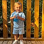 toddler, child, smiling, outdoor, wooden_deck, greenery, sunlight, blue_shirt, shorts, sneakers, sippy_cup, happy, standing, daylight, wooden_railing, casual_clothing, person, face, legs, feet