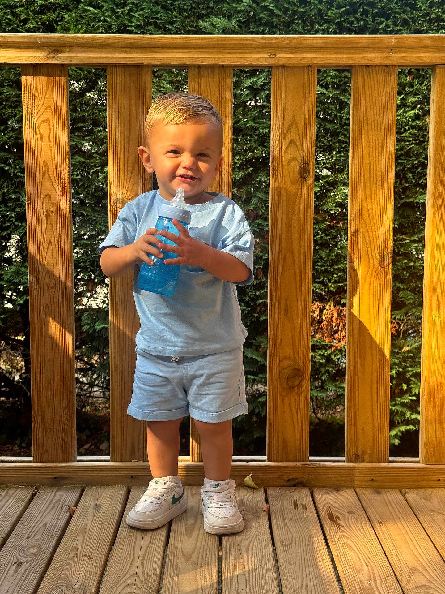 Liam participe au concours pour gagner de l'argent avec cette photo : toddler, child, smiling, outdoor, wooden_deck, greenery, sunlight, blue_shirt, shorts, sneakers, sippy_cup, happy, standing, daylight, wooden_railing, casual_clothing, person, face, legs, feet