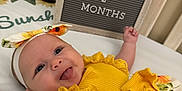 Lainey is registered to the contest to win money with this photo: baby, infant, smiling, tongue_out, bed, headband, yellow_clothing, sunflower_pattern, letter_board, text, milestone, cute, happy, portrait, child, indoor, person, fabric, playful, baby_girl